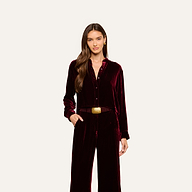 A woman with long brown hair wears the Cosette Velvet Shirt in burgundy along with matching velvet pants, accessorized with a brown belt featuring a square gold buckle, standing against a plain light background.
