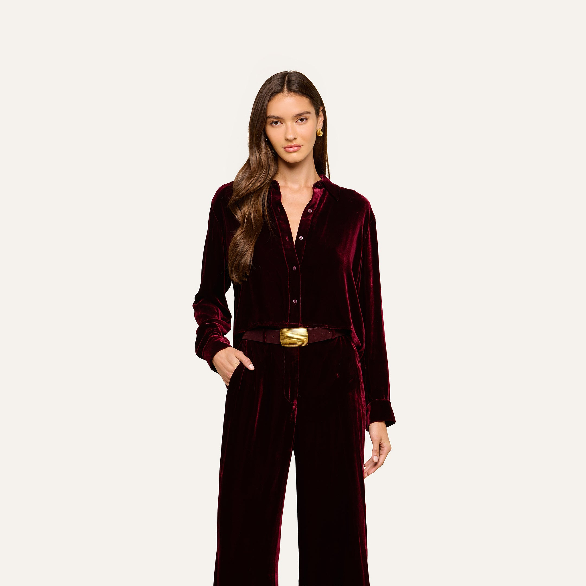 A woman with long brown hair wears the Cosette Velvet Shirt in burgundy along with matching velvet pants, accessorized with a brown belt featuring a square gold buckle, standing against a plain light background.