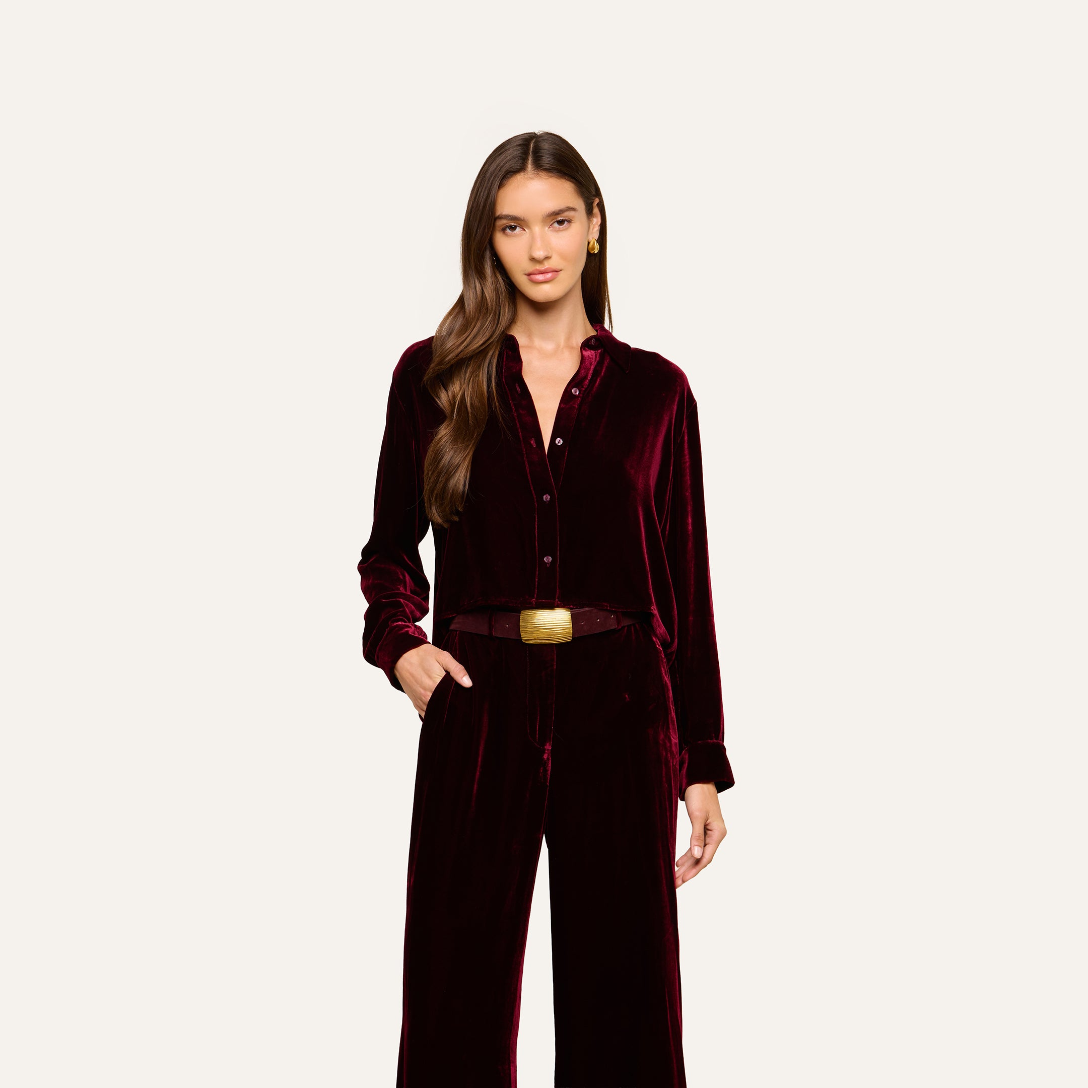 A woman with long brown hair wears the Cosette Velvet Shirt in burgundy along with matching velvet pants, accessorized with a brown belt featuring a square gold buckle, standing against a plain light background.