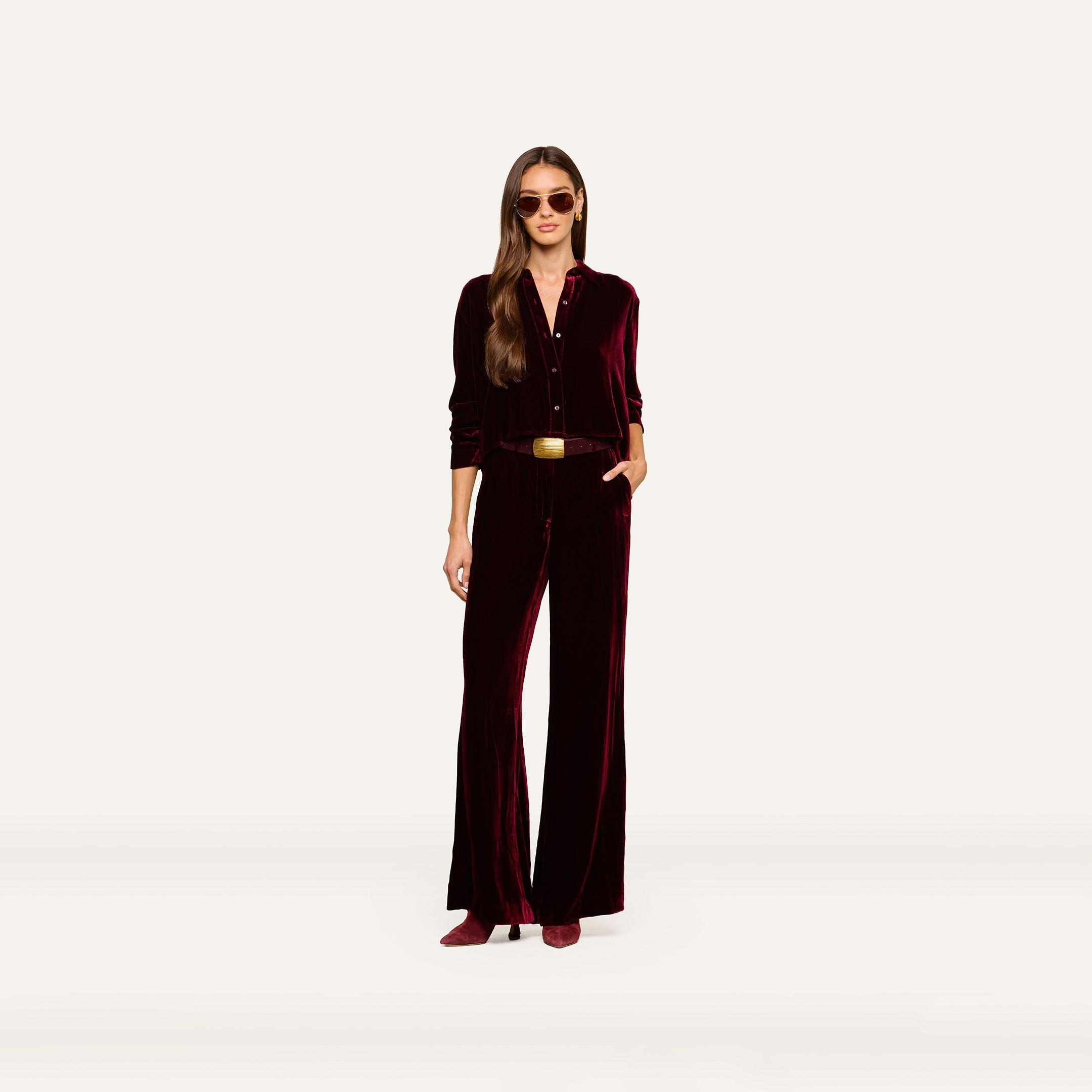 A woman stands against a plain white background with one hand in her pocket, wearing the Cosette Velvet Shirt styled as a jumpsuit, along with matching shoes, sunglasses, and a belt.