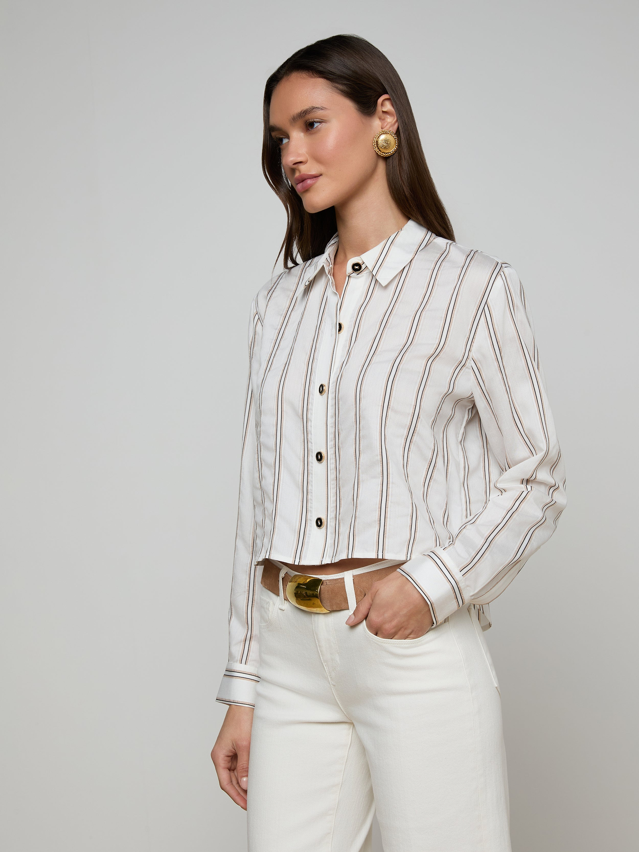 A woman wears the Cosette Shirt—a white button-up tailored shirt with brown jacquard stripes—paired with white pants, a tan belt, and large round earrings, standing against a plain light background.