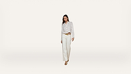 A woman stands against a plain white background, wearing a striped button-down shirt tucked into high-waisted white pants with a tan belt and beige heels.