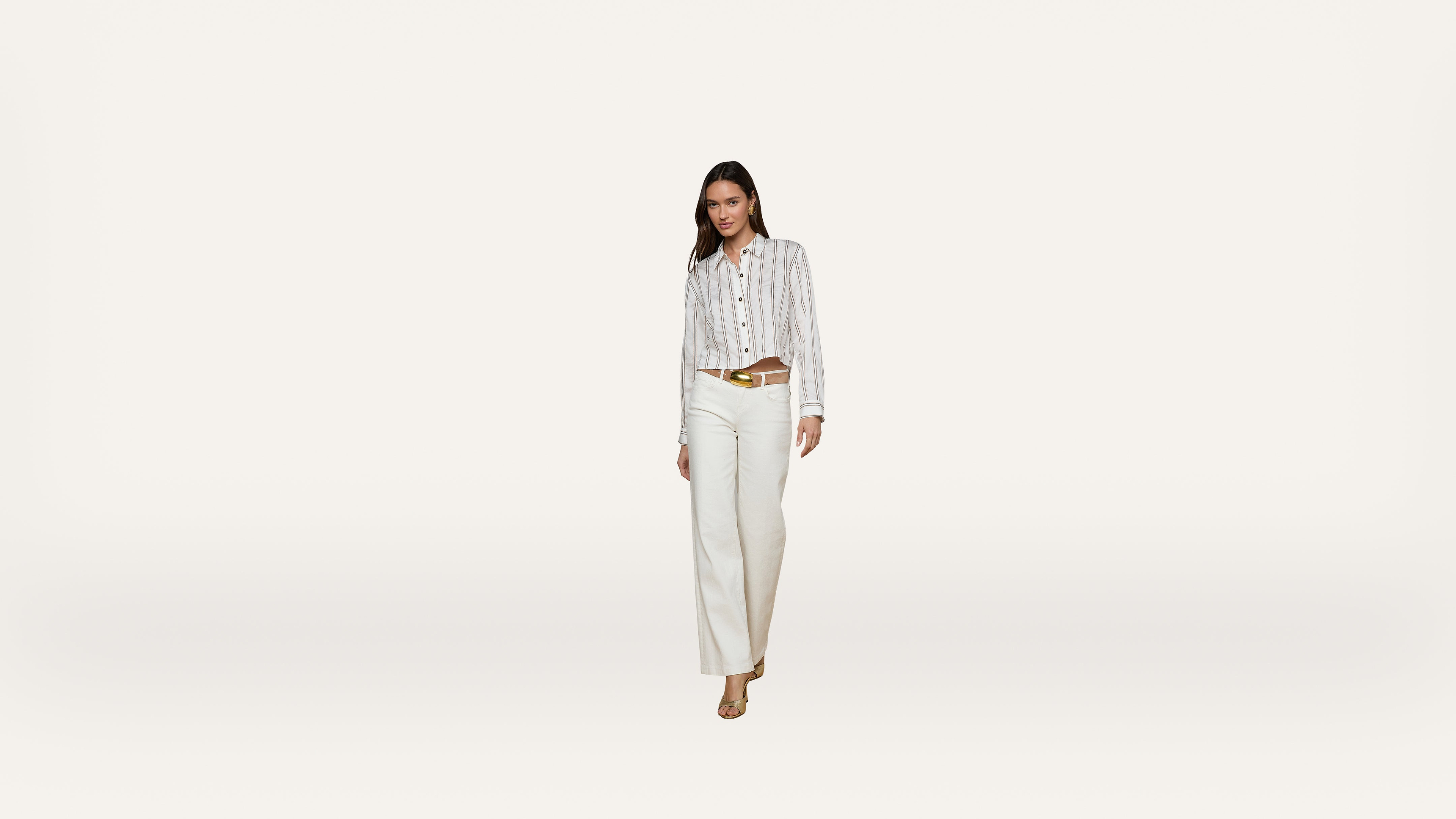 A woman stands against a plain white background, wearing a striped button-down shirt tucked into high-waisted white pants with a tan belt and beige heels.