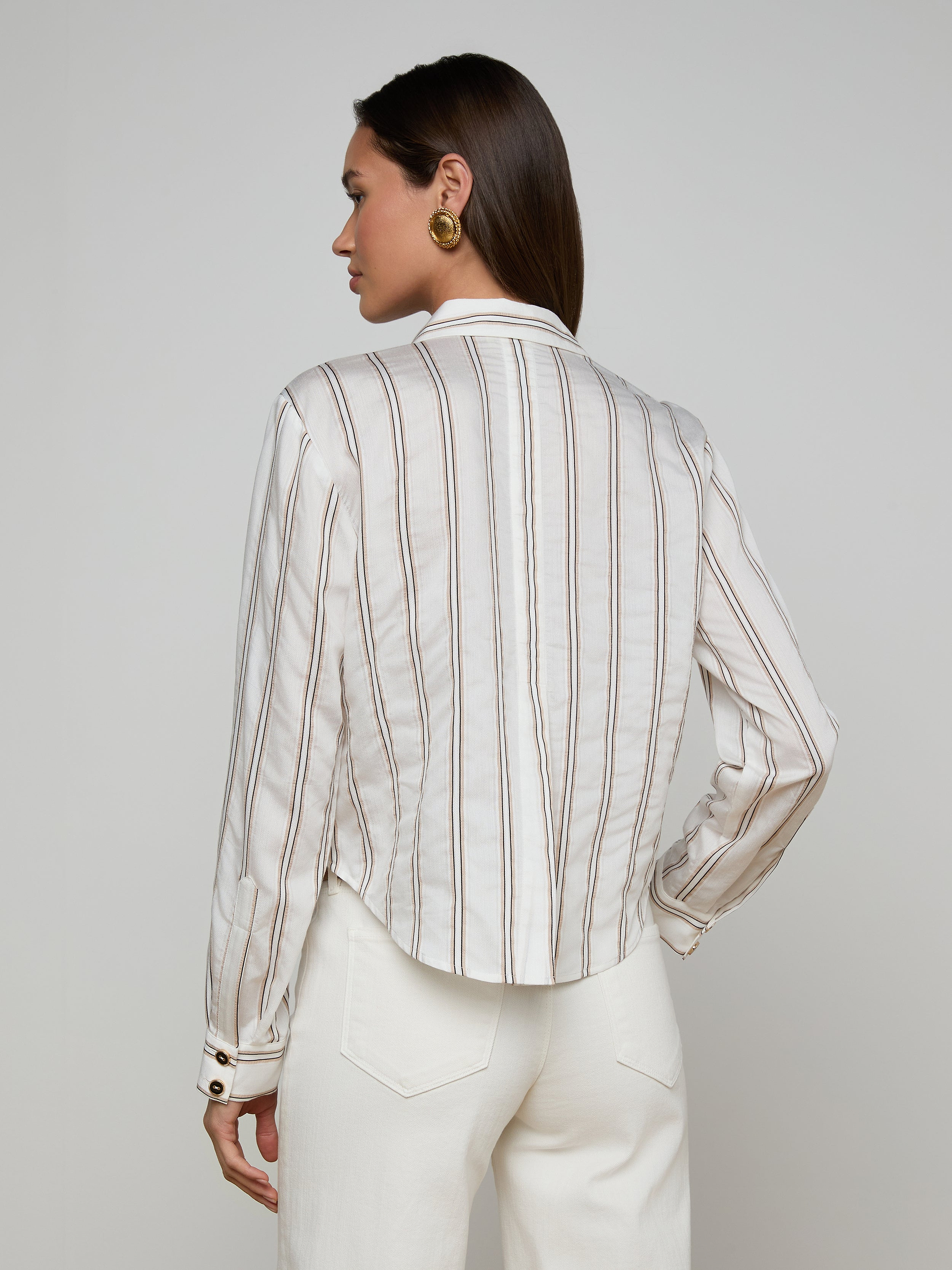 A woman stands facing away, wearing the Cosette Shirt—a tailored white long-sleeve top with vertical jacquard stripes—paired with white pants. She has straight brown hair and accessorizes with gold round earrings, set against a plain, light-colored background.