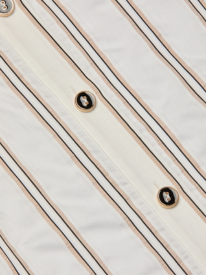 A close-up of the Cosette Shirt’s jacquard white fabric with vertical beige and black stripes, detailed with round gold-rimmed, black-centered buttons sewn along its cropped high-low hem.
