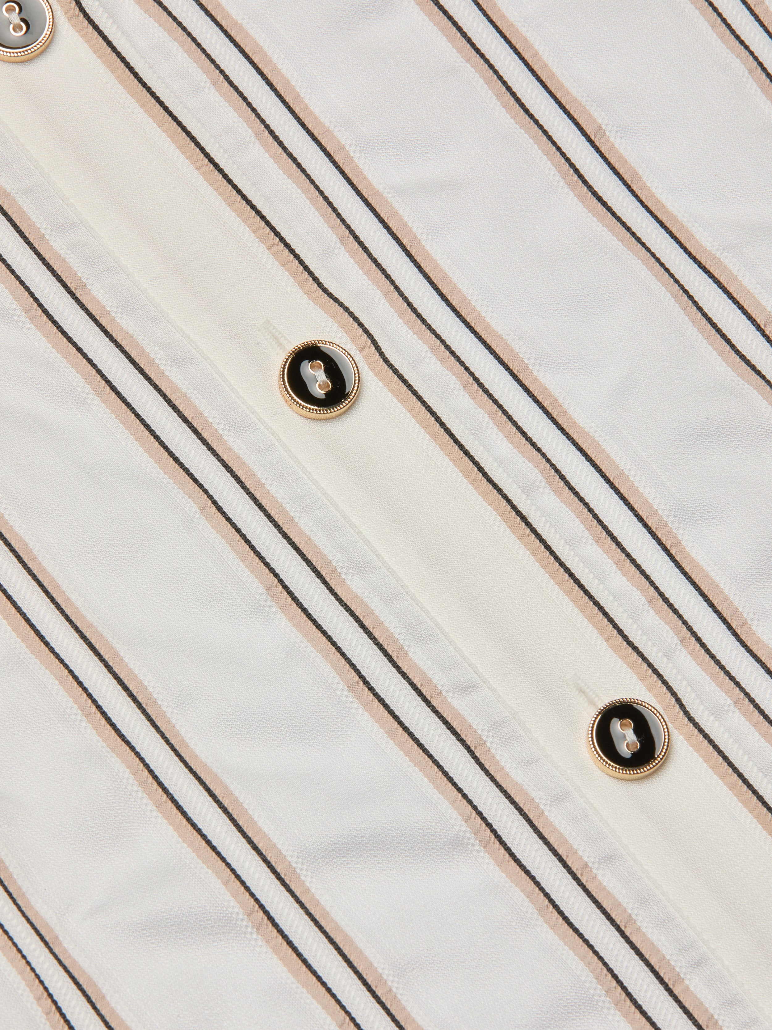 Close-up of the Cosette Shirt, crafted from white fabric with vertical beige and black jacquard stripes and accented by gold-rimmed black buttons sewn along the stripes, evoking a tailored look.