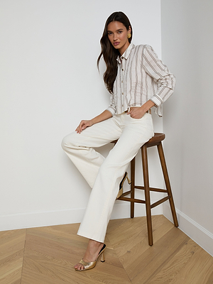 A woman with long brown hair sits on a wooden stool in a corner, wearing the Cosette Shirt, white wide-leg pants, and gold heeled sandals. She poses with one hand in her pocket and a relaxed expression.