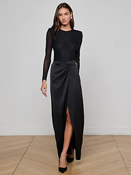 A woman wearing the Trista Bodysuit—with long sleeves, structured shoulder pads, and a thigh-high slit—paired with black heels, stands against a plain white background.