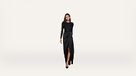 A woman in a long-sleeve black dress with a thigh-high slit stands against a plain white background, looking forward with a neutral expression.