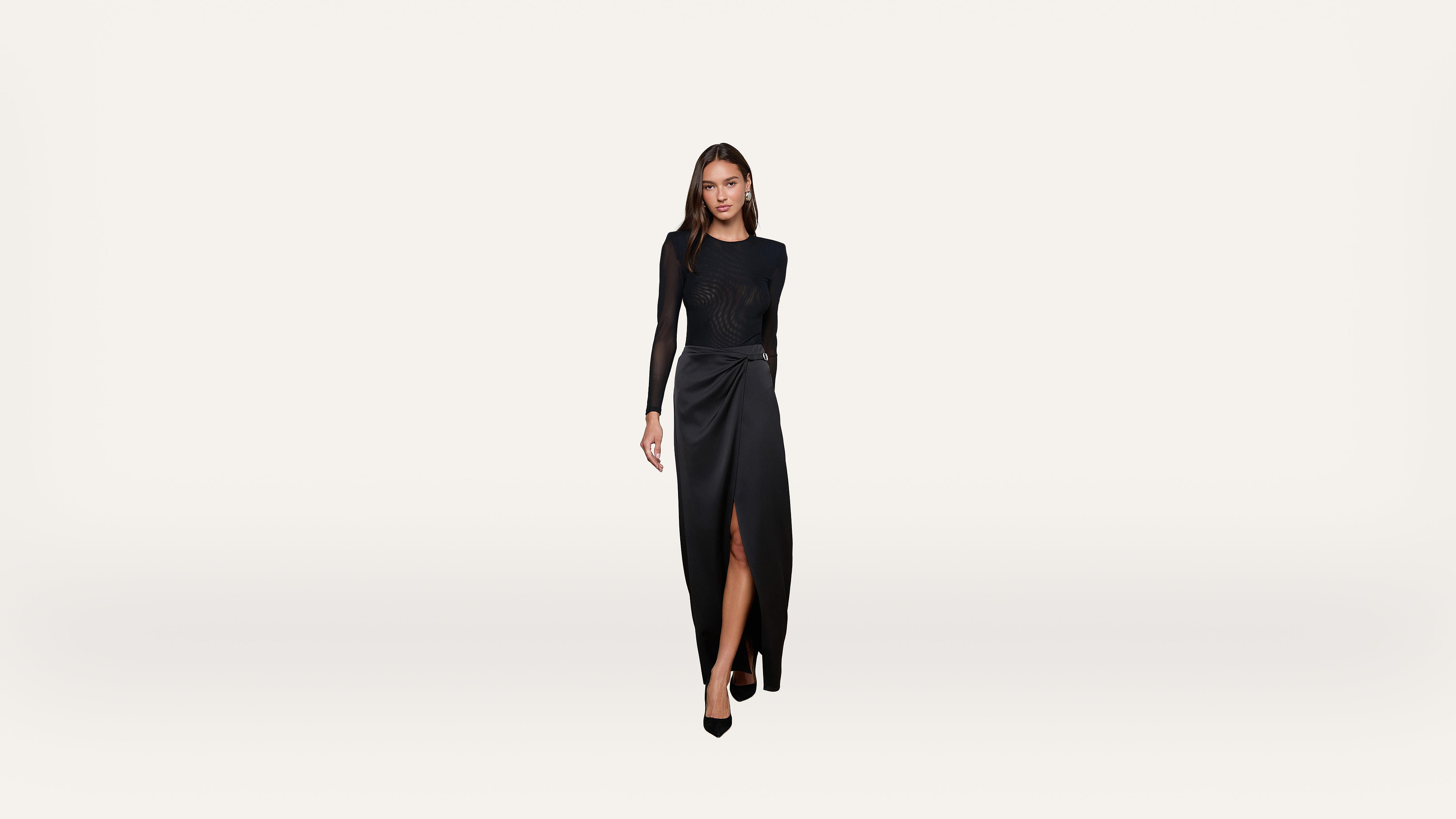A woman in a long-sleeve black dress with a thigh-high slit stands against a plain white background, looking forward with a neutral expression.