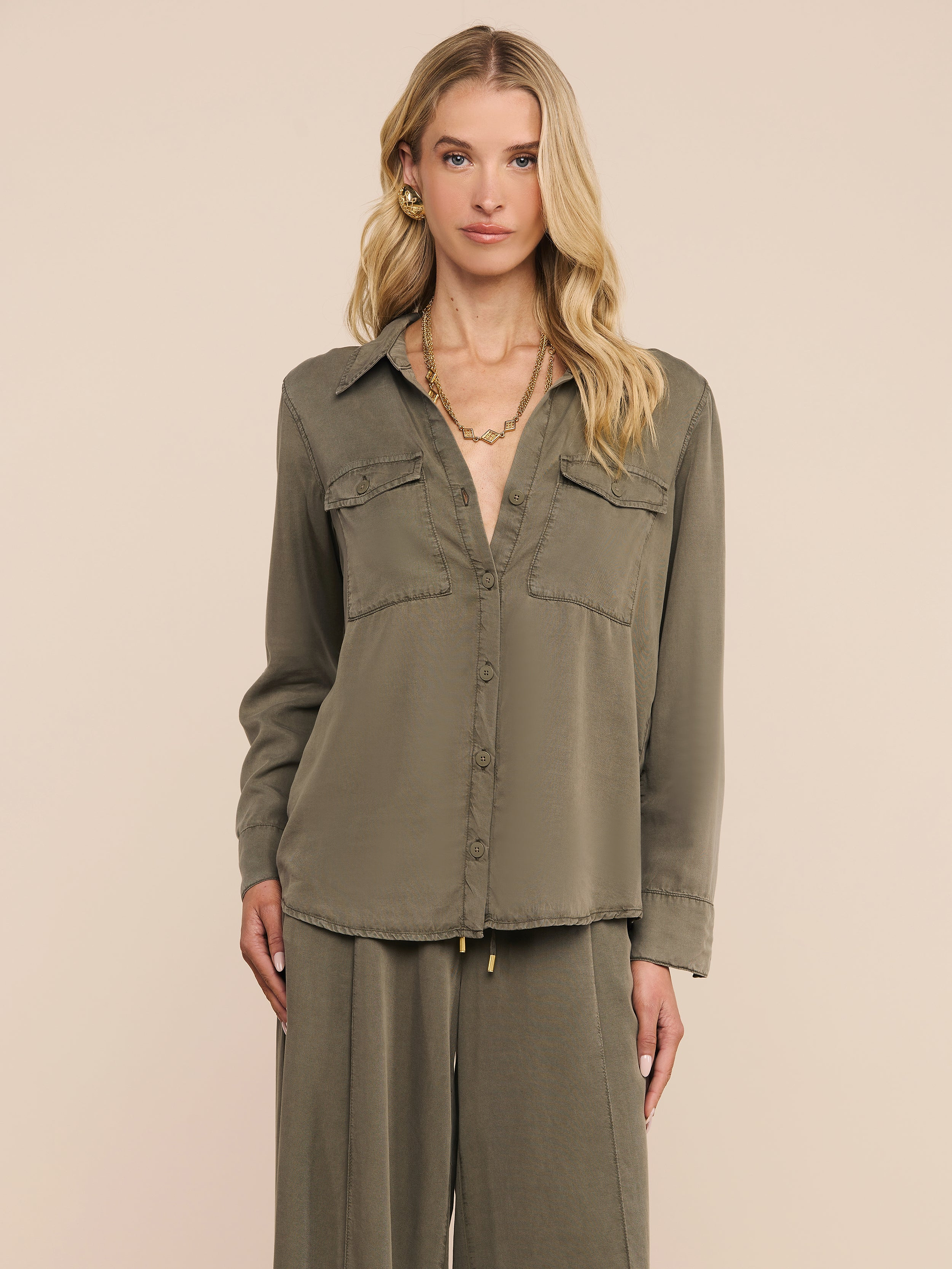 A woman with long blonde hair wears the Pearson Shirt—a smoky olive green button-down—in lightweight lyocell, paired with matching wide-leg pants, standing against a plain light background. She accessorizes with gold hoop earrings and a gold necklace.