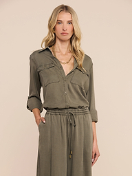 Woman with long blonde hair wearing the Pearson Shirt in smoky olive green lyocell, styled with rolled sleeves and a drawstring waist, standing with one hand in her pocket against a plain light background.