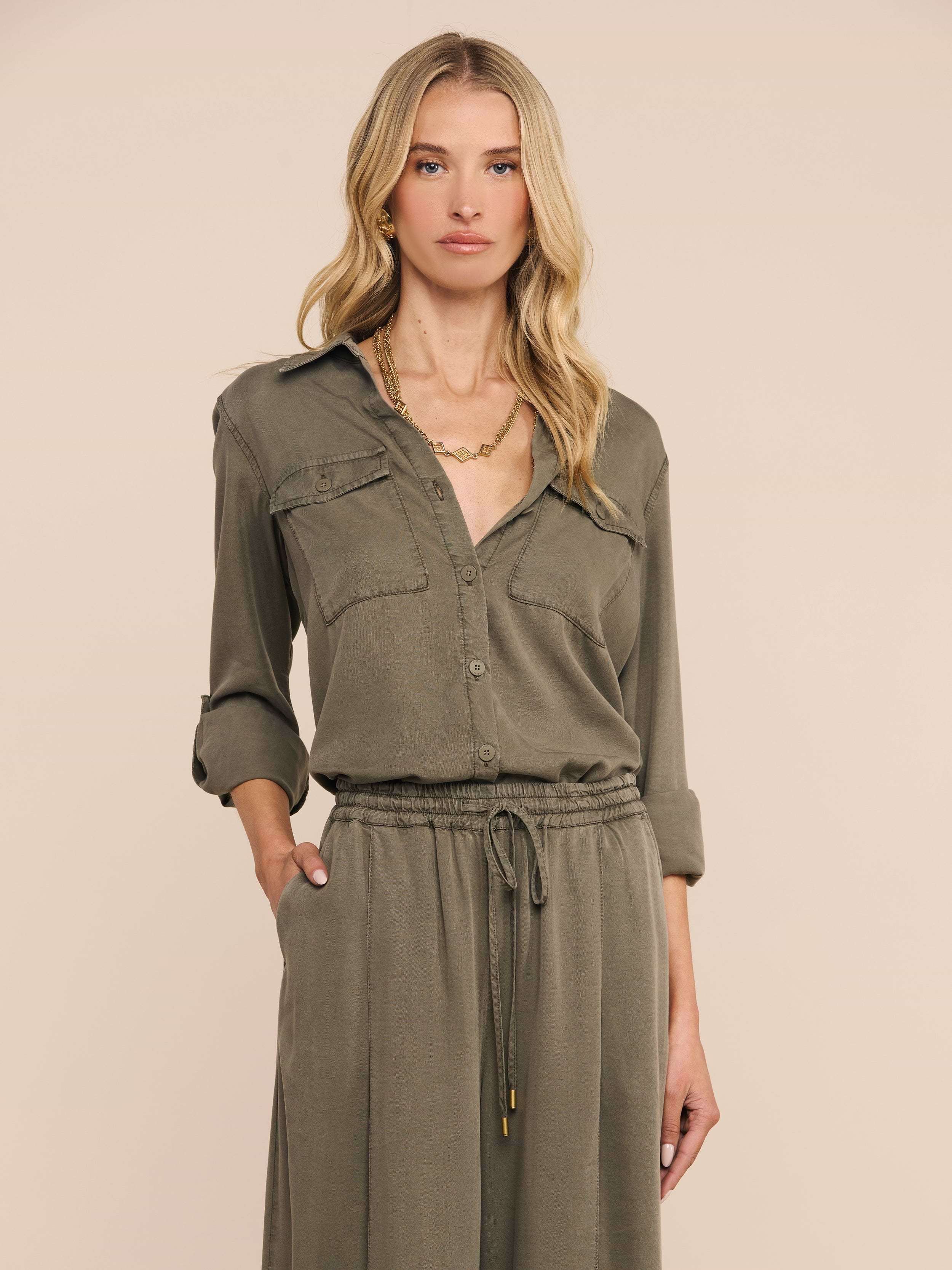 Woman with long blonde hair wearing the Pearson Shirt in smoky olive green lyocell, styled with rolled sleeves and a drawstring waist, standing with one hand in her pocket against a plain light background.