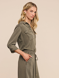 Woman with wavy blonde hair wearing the Pearson Shirt—a lightweight lyocell, button-down shirt dress in smoky olive green with side pockets—standing with one hand in a pocket against a plain light background.