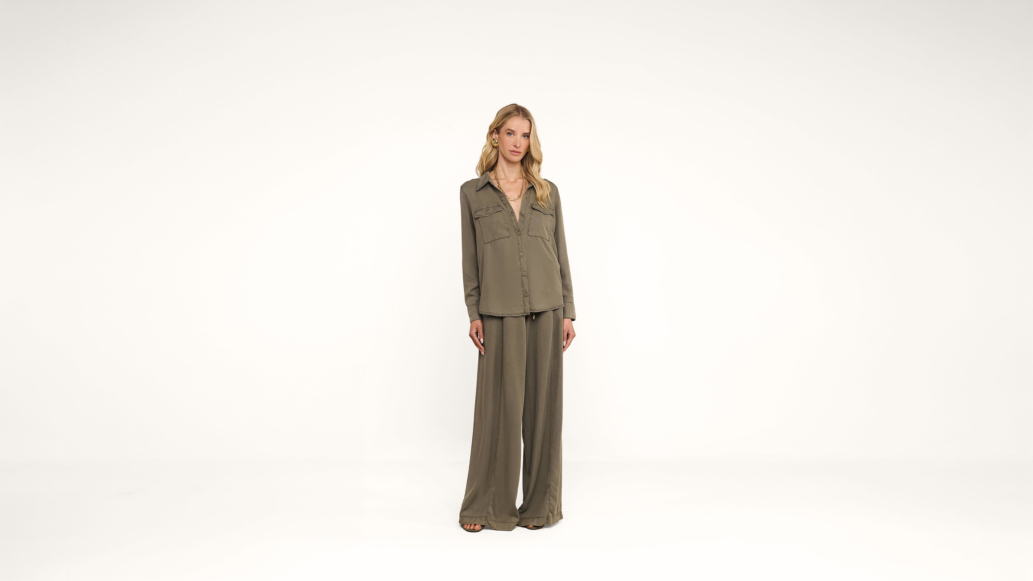 A person stands in the center of a white background wearing an olive green long-sleeved shirt and matching wide-leg pants.