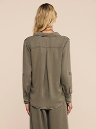 A woman with wavy blonde hair is shown from the back against a plain light background, wearing the Pearson Shirt in a smoky olive green, paired with matching loose pants.