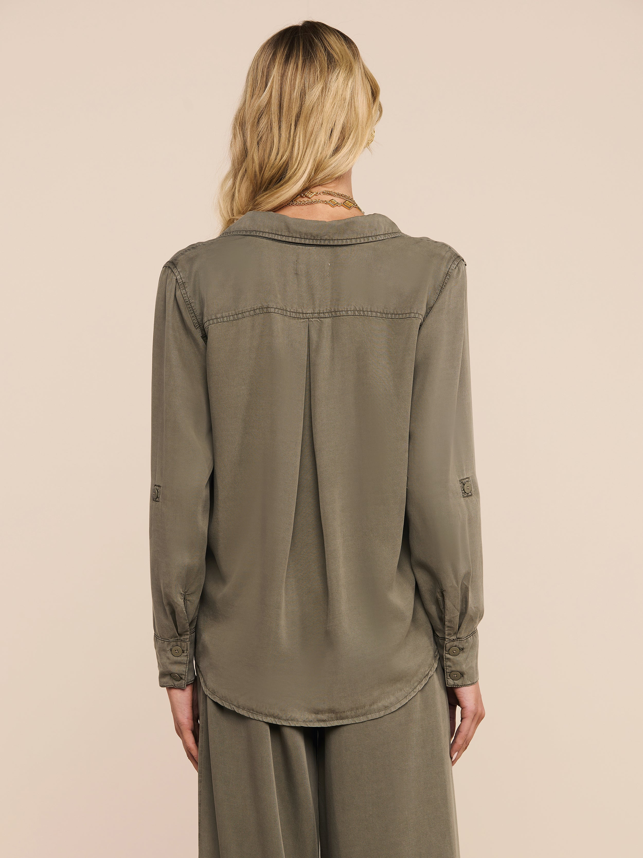 A woman with wavy blonde hair is shown from the back against a plain light background, wearing the Pearson Shirt in a smoky olive green, paired with matching loose pants.