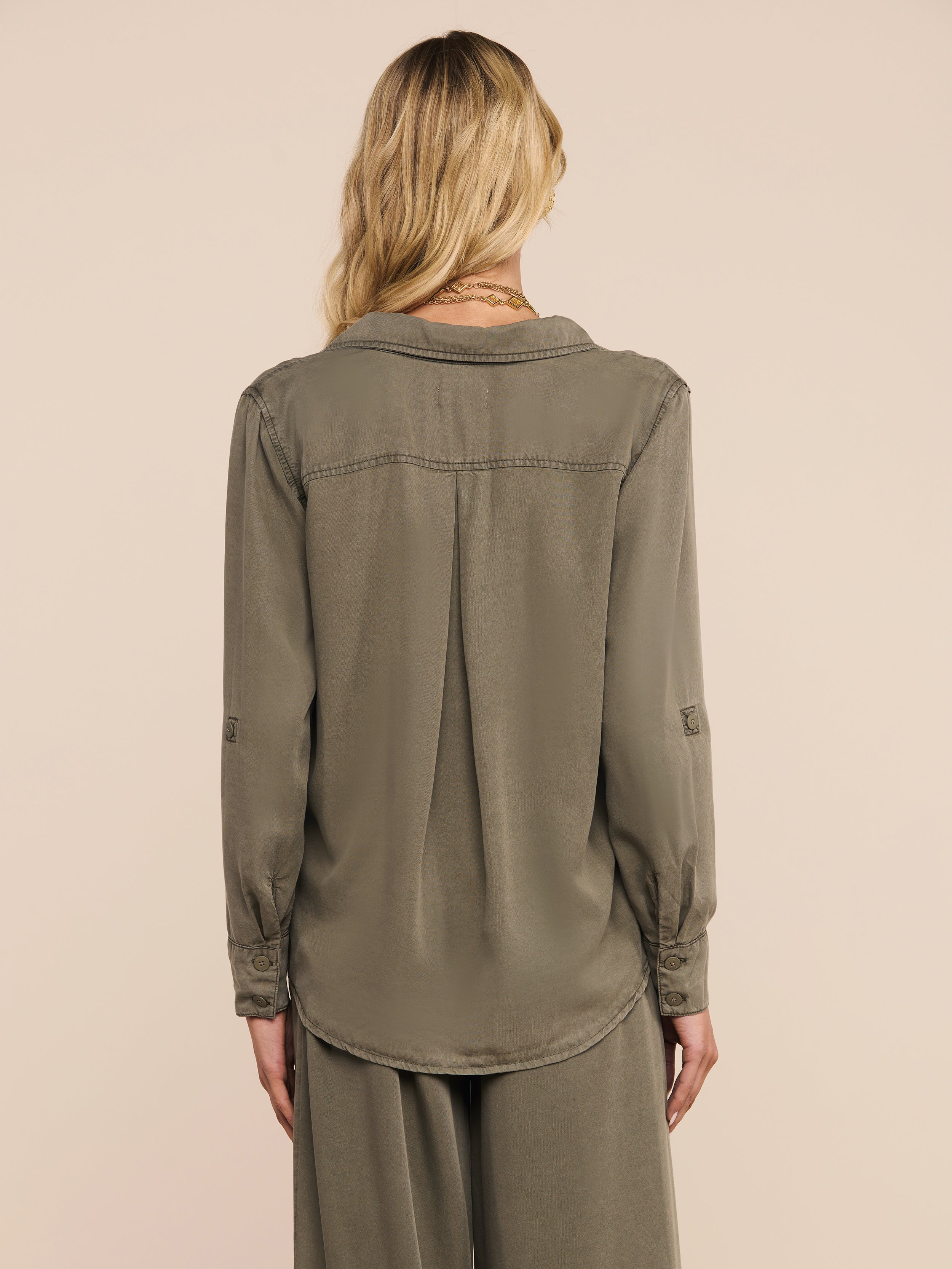 A woman with wavy blonde hair is shown from the back against a plain light background, wearing the Pearson Shirt in a smoky olive green, paired with matching loose pants.