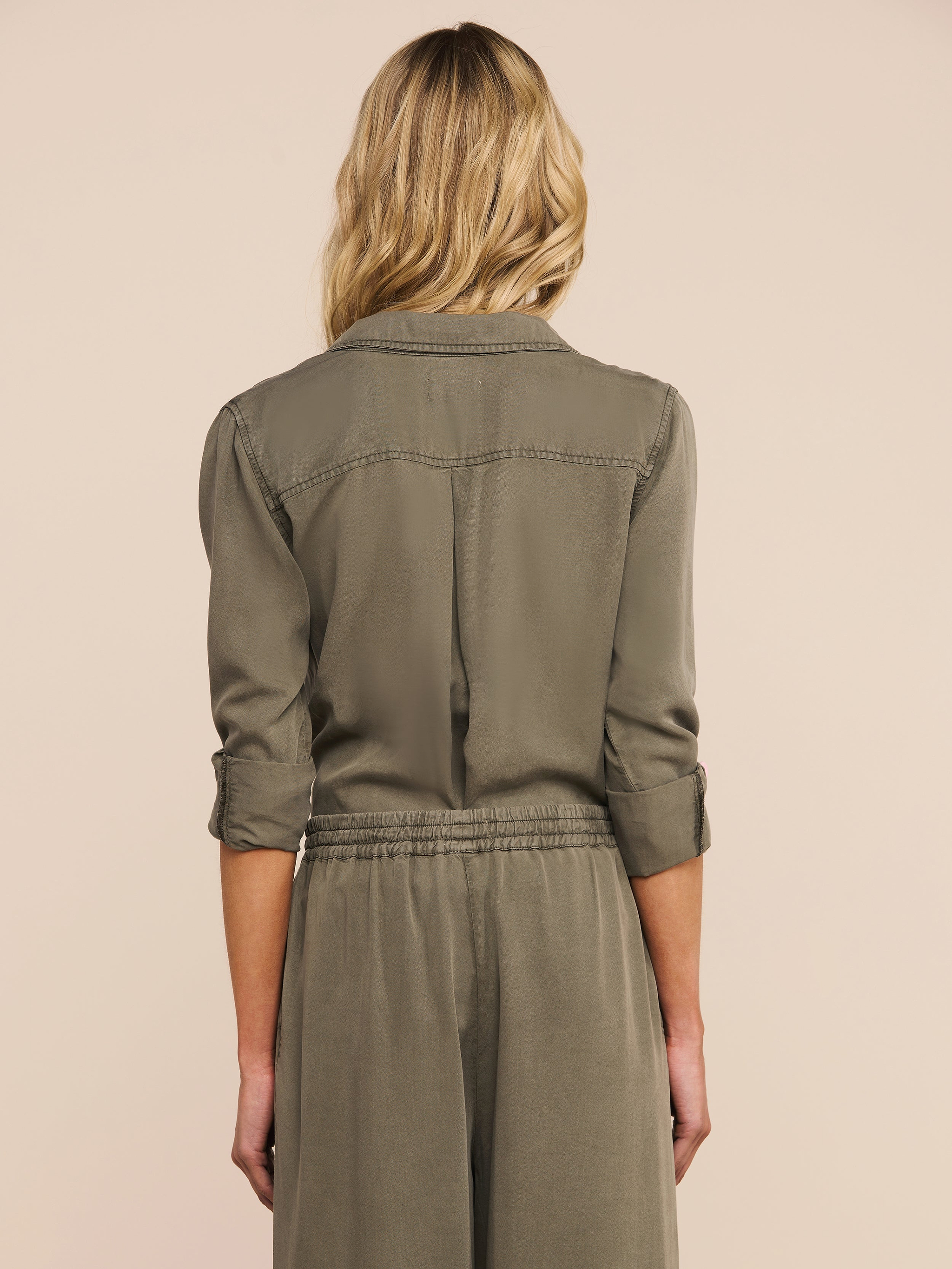 A person with wavy blonde hair, wearing the Pearson Shirt in a smoky olive green lightweight lyocell fabric, is standing with their back to the camera against a plain, light background.