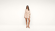 A woman wearing sunglasses and a light beige, long-sleeve, belted romper stands confidently in high heels against a plain white background.