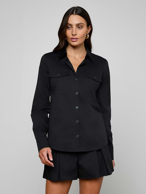 A woman stands against a plain background wearing the Pearson Poplin Shirt in black, featuring two chest pockets and matching black shorts.
