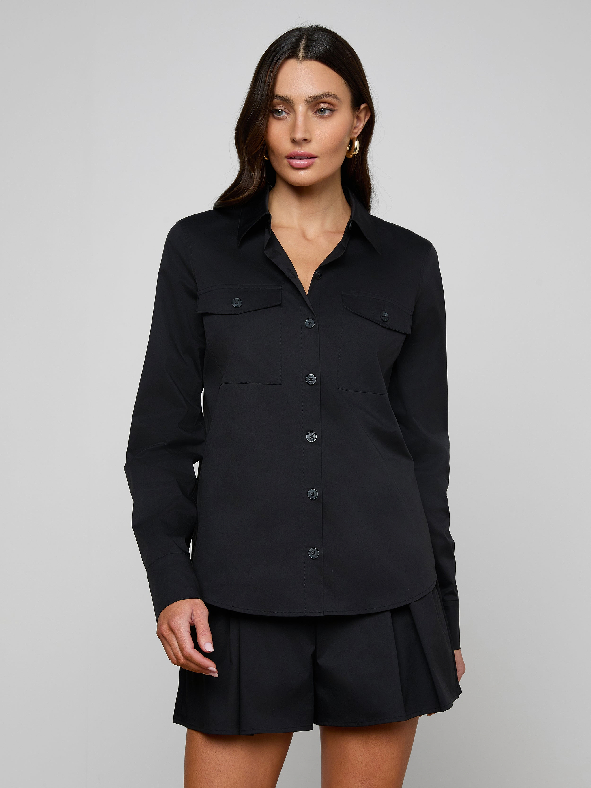 A woman stands against a plain background wearing the Pearson Poplin Shirt in black, featuring two chest pockets and matching black shorts.