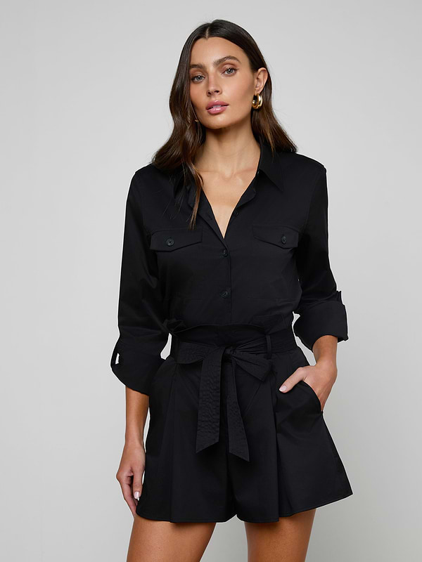The woman is wearing the Pearson Poplin Shirt in black, styled with a belted waist and buttoned chest pockets, resembling a romper with long sleeves, as she stands with one hand in her pocket against a plain light background.