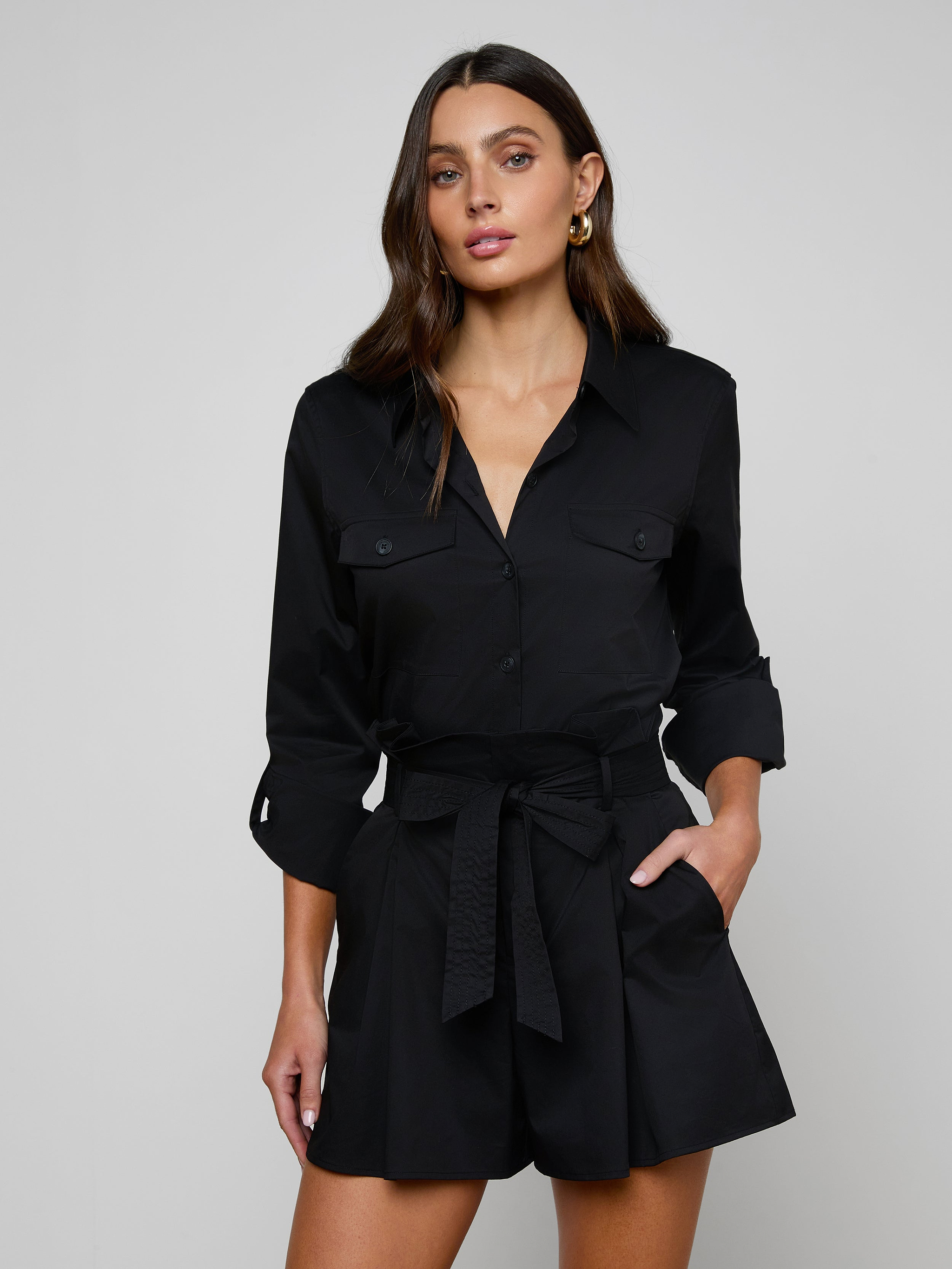 The woman is wearing the Pearson Poplin Shirt in black, styled with a belted waist and buttoned chest pockets, resembling a romper with long sleeves, as she stands with one hand in her pocket against a plain light background.
