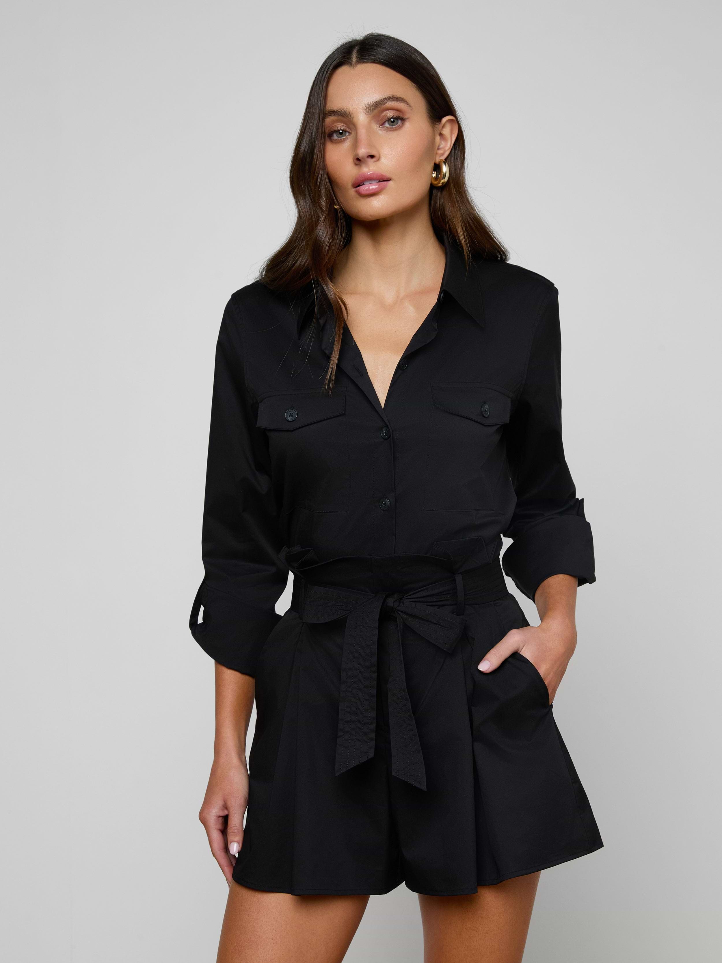 The woman is wearing the Pearson Poplin Shirt in black, styled with a belted waist and buttoned chest pockets, resembling a romper with long sleeves, as she stands with one hand in her pocket against a plain light background.