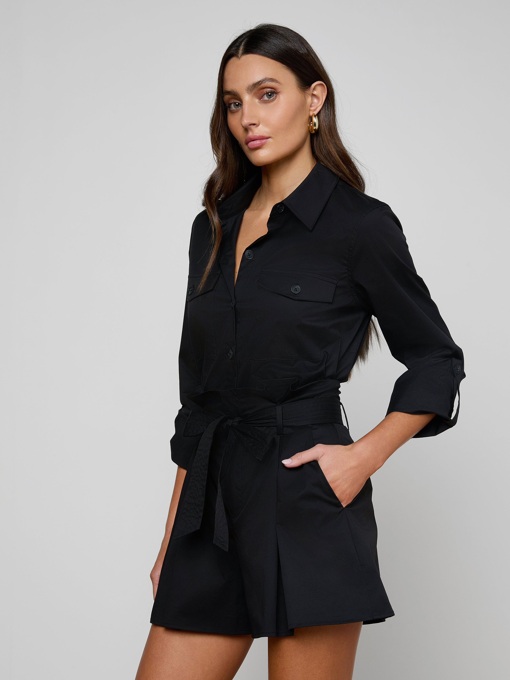 A woman wearing the Pearson Poplin Shirt, a black button-down dress with rolled-up long sleeves, stands with one hand in her pocket against a plain, light background.