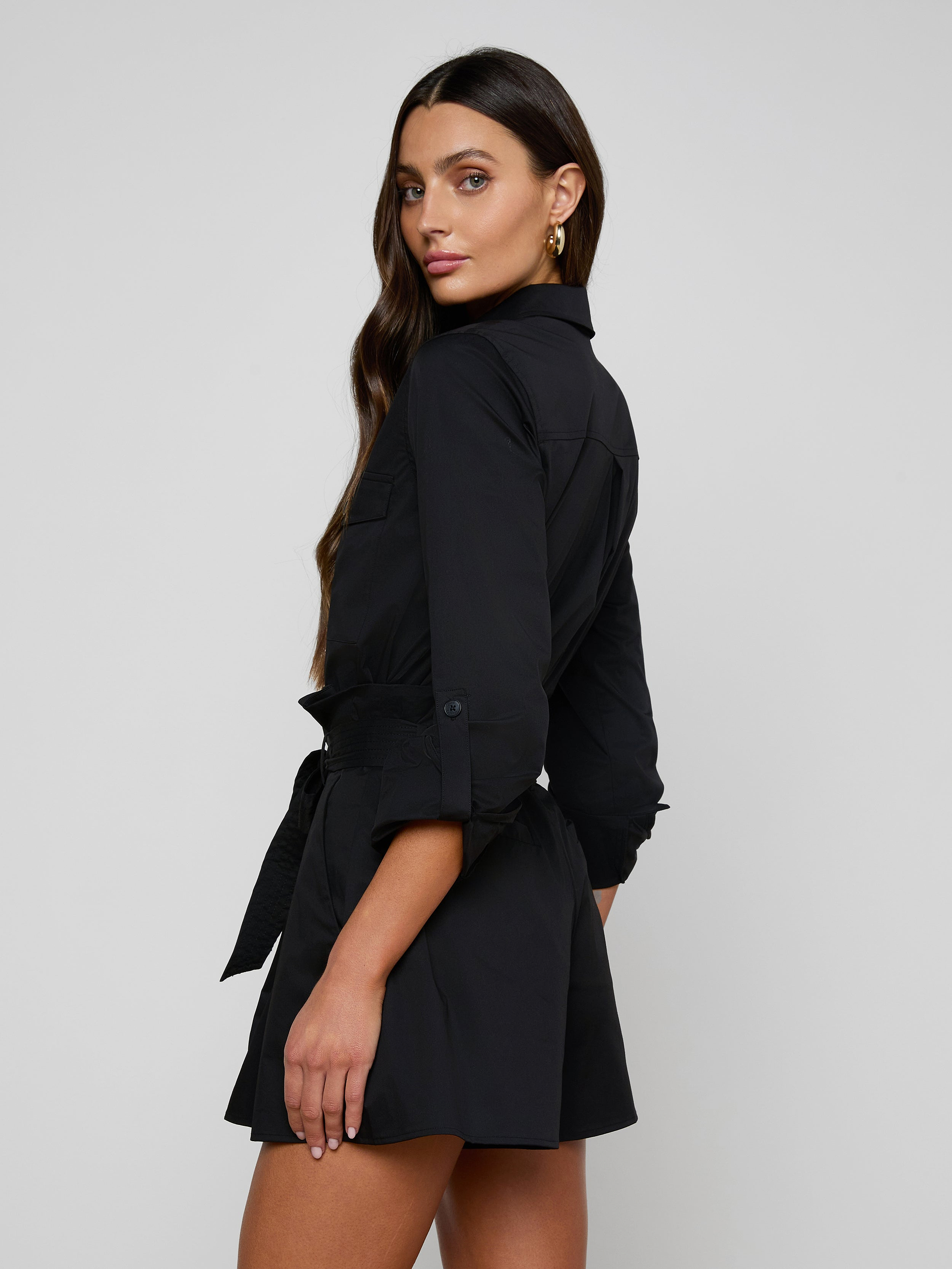 A woman wearing the Pearson Poplin Shirt, styled with long sleeves and a belted waist to resemble a button-down romper, stands slightly turned to the side, looking at the camera against a plain light background.