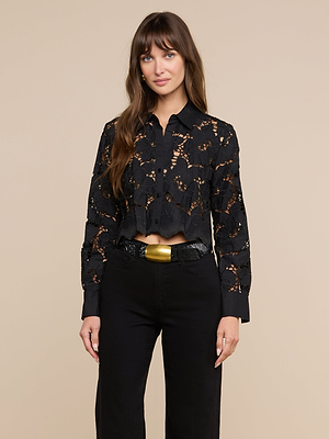 A woman with long brown hair and bangs stands against a beige background, wearing the Seychelle Cropped Eyelet Blouse in black, paired with black pants and a black belt with a large gold buckle. She has a relaxed expression.
