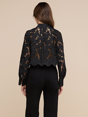 تظهر امرأة ذات شعر بني طويل من الخلف، وهي ترتدي بلوزة «Seychelle Cropped Eyelet Blouse» — وهي بلوزة سوداء من الدانتيل بنقوش زهرية وتفاصيل شفافة — وسروالاً أسود على خلفية بيج.