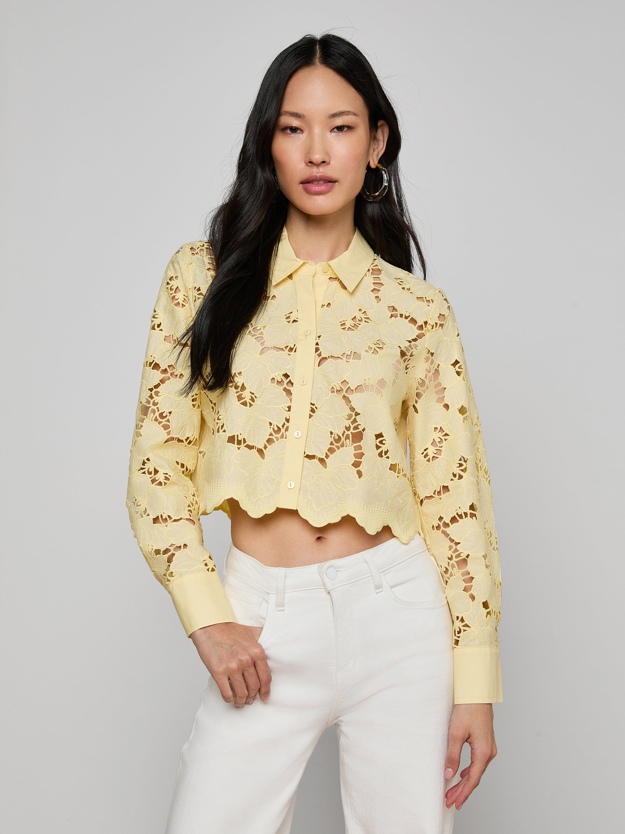 A woman wearing the Seychelle Cropped Eyelet Blouse—a light yellow, long-sleeve poplin lace crop top with a scalloped hem—paired with high-waisted white pants, stands against a plain background.