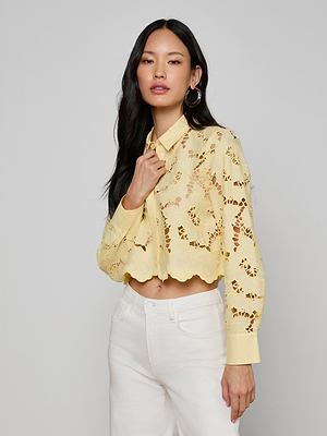 A woman with long dark hair stands against a plain background, wearing the Seychelle Cropped Eyelet Blouse in pale yellow with floral cutouts and white high-waisted pants. She has one hand near her collar and a neutral expression.