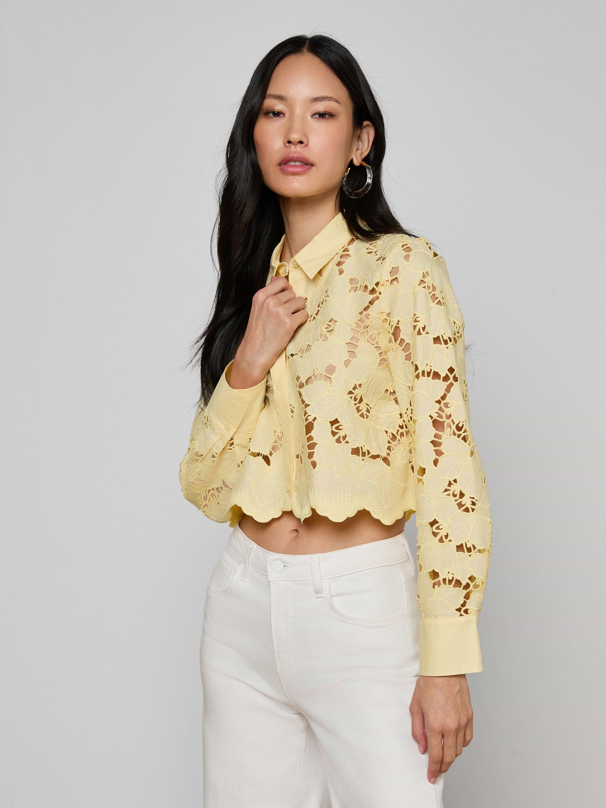 A woman in the Seychelle Cropped Eyelet Blouse—a pale yellow, floral eyelet top with long sleeves—paired with white pants stands against a plain background, looking confidently at the camera.