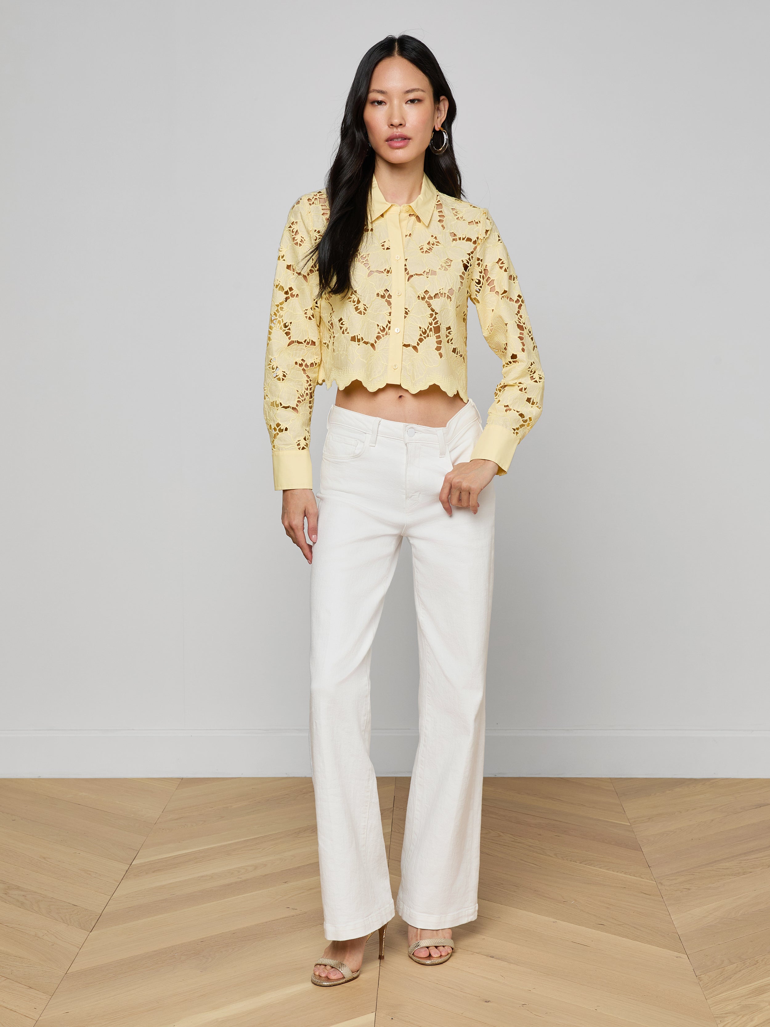 A woman stands against a plain white background wearing the Seychelle Cropped Eyelet Blouse, a yellow long-sleeve eyelet top, paired with white wide-leg pants and heeled sandals.