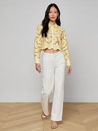 A woman walks forward wearing the Seychelle Cropped Eyelet Blouse, featuring a yellow floral eyelet design, paired with white wide-leg pants and open-toe shoes, against a plain light background.