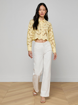 A woman with long dark hair wears the Seychelle Cropped Eyelet Blouse and high-waisted white pants, walking indoors on a light wood floor against a plain light gray wall.