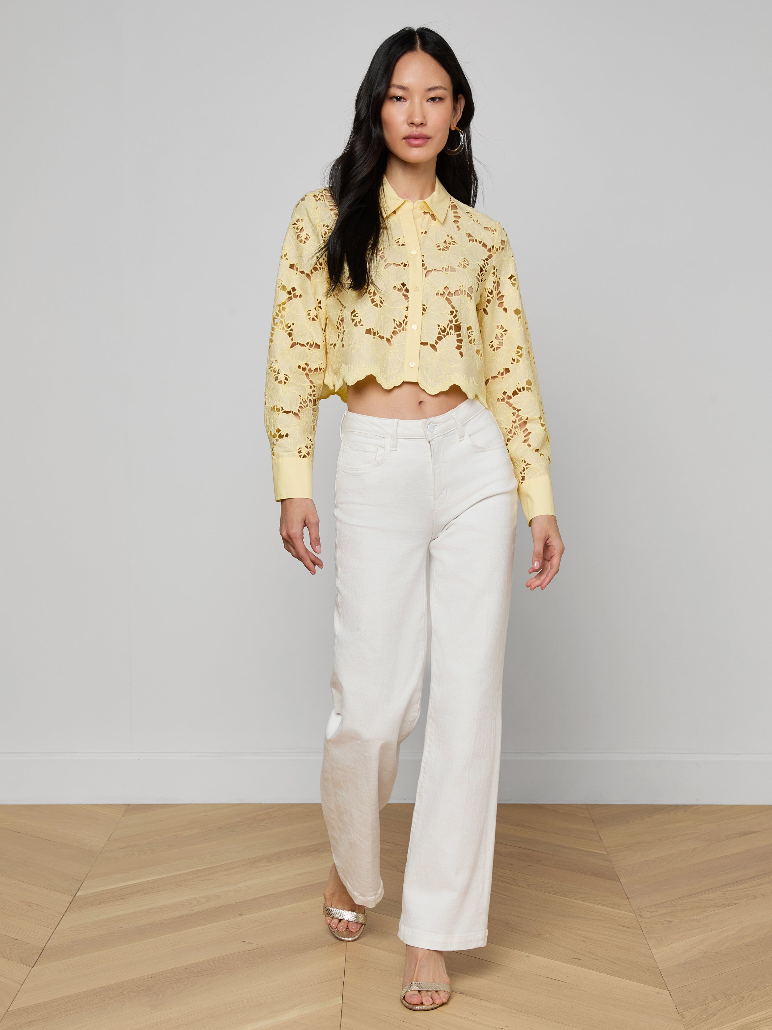 A woman walks forward wearing the Seychelle Cropped Eyelet Blouse, featuring a yellow floral eyelet design, paired with white wide-leg pants and open-toe shoes, against a plain light background.