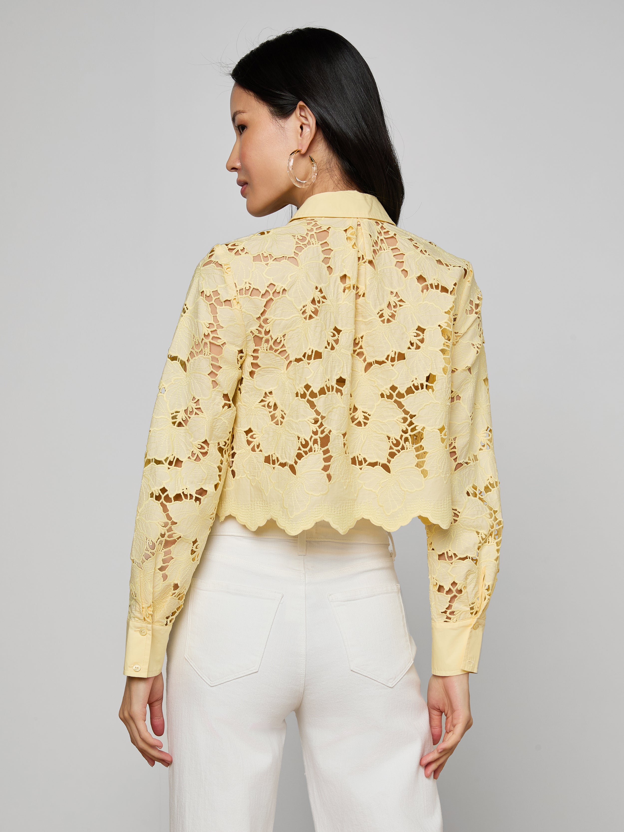 A woman with long dark hair, wearing large hoop earrings and high-waisted white pants, is shown from the back against a plain light background, modeling the Seychelle Cropped Eyelet Blouse in yellow sheer floral fabric.