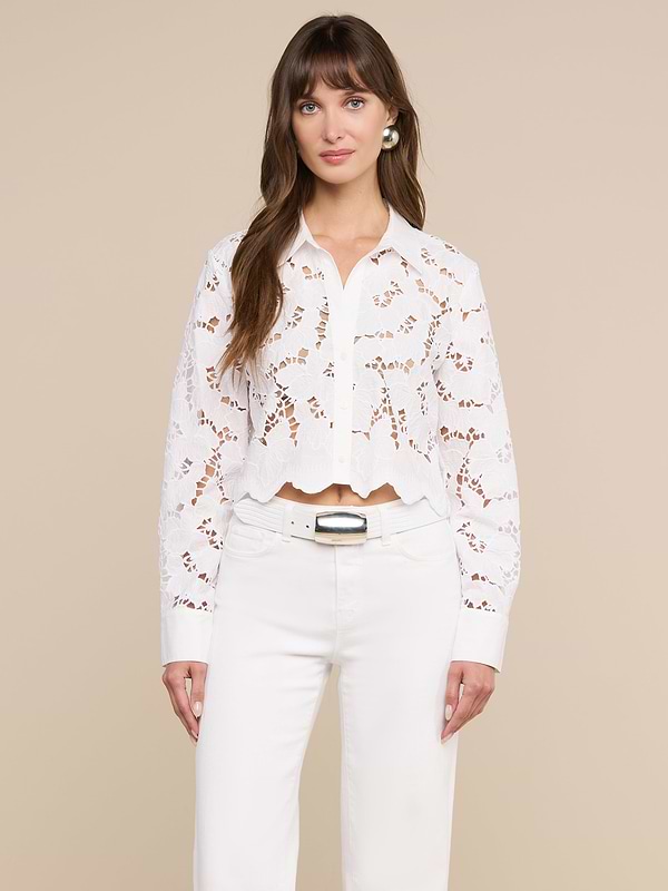 A woman is wearing the Seychelle Cropped Eyelet Blouse—a long-sleeved, white floral lace embroidered top with scalloped edges—paired with high-waisted white belted pants, standing against a plain light background.