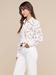 A woman with long brown hair stands in profile against a plain light background, wearing the Seychelle Cropped Eyelet Blouse paired with high-waisted white pants.