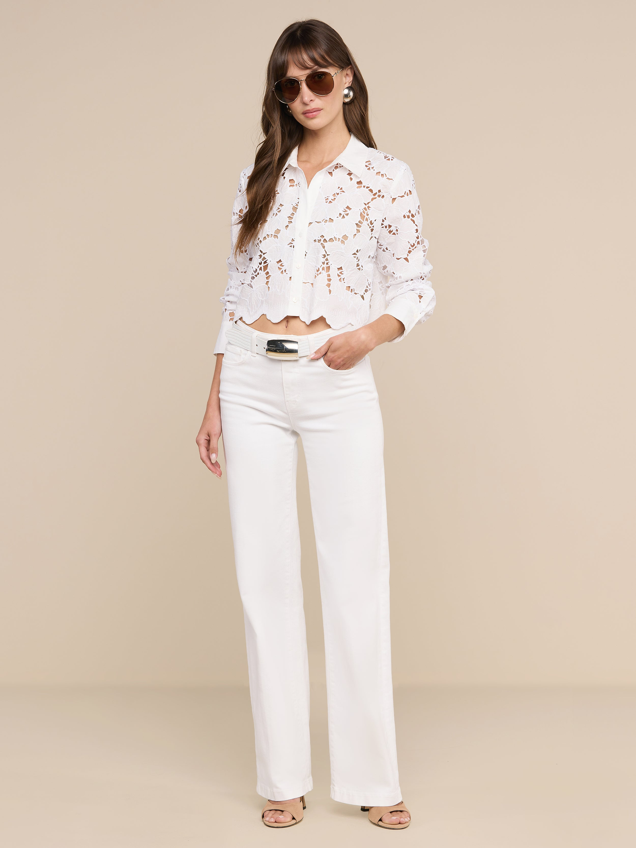 A woman wearing sunglasses and the Seychelle Cropped Eyelet Blouse— a white long-sleeve lace top with red pattern detailing— paired with white wide-leg pants and beige sandals, stands against a plain white background, effortlessly capturing relaxed beach style.