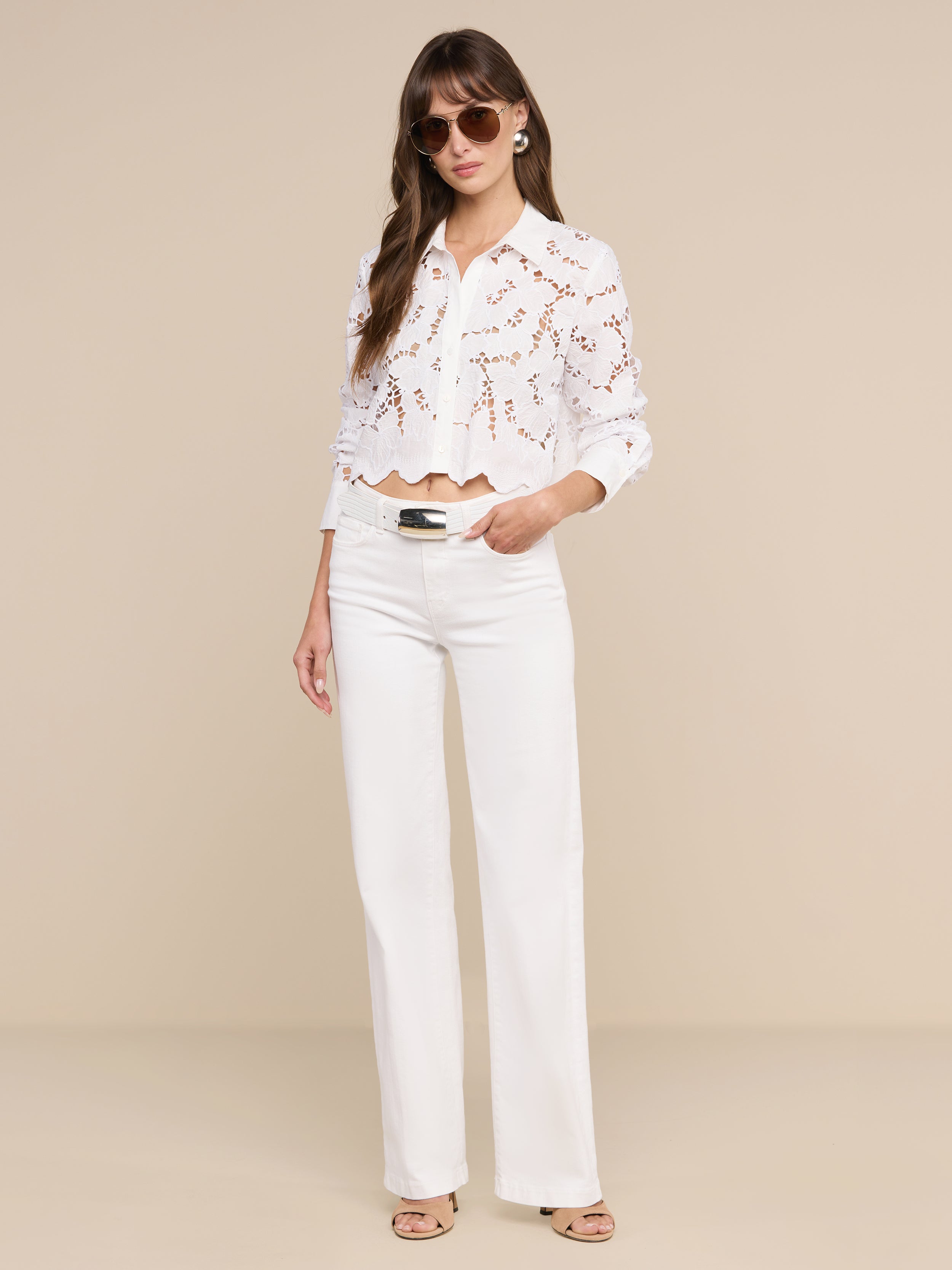A woman wearing sunglasses and the Seychelle Cropped Eyelet Blouse— a white long-sleeve lace top with red pattern detailing— paired with white wide-leg pants and beige sandals, stands against a plain white background, effortlessly capturing relaxed beach style.
