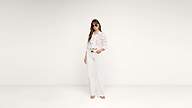 Woman wearing sunglasses, a white blouse with a floral pattern, and white wide-leg pants stands against a plain white background.
