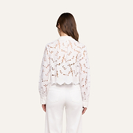 A woman stands with her back to the camera, wearing the Seychelle Cropped Eyelet Blouse—an all-white, long-sleeve top featuring a semi-sheer floral eyelet pattern and scalloped hem—paired with high-waisted white pants. Her brown hair is worn down, and the background is a plain light beige.