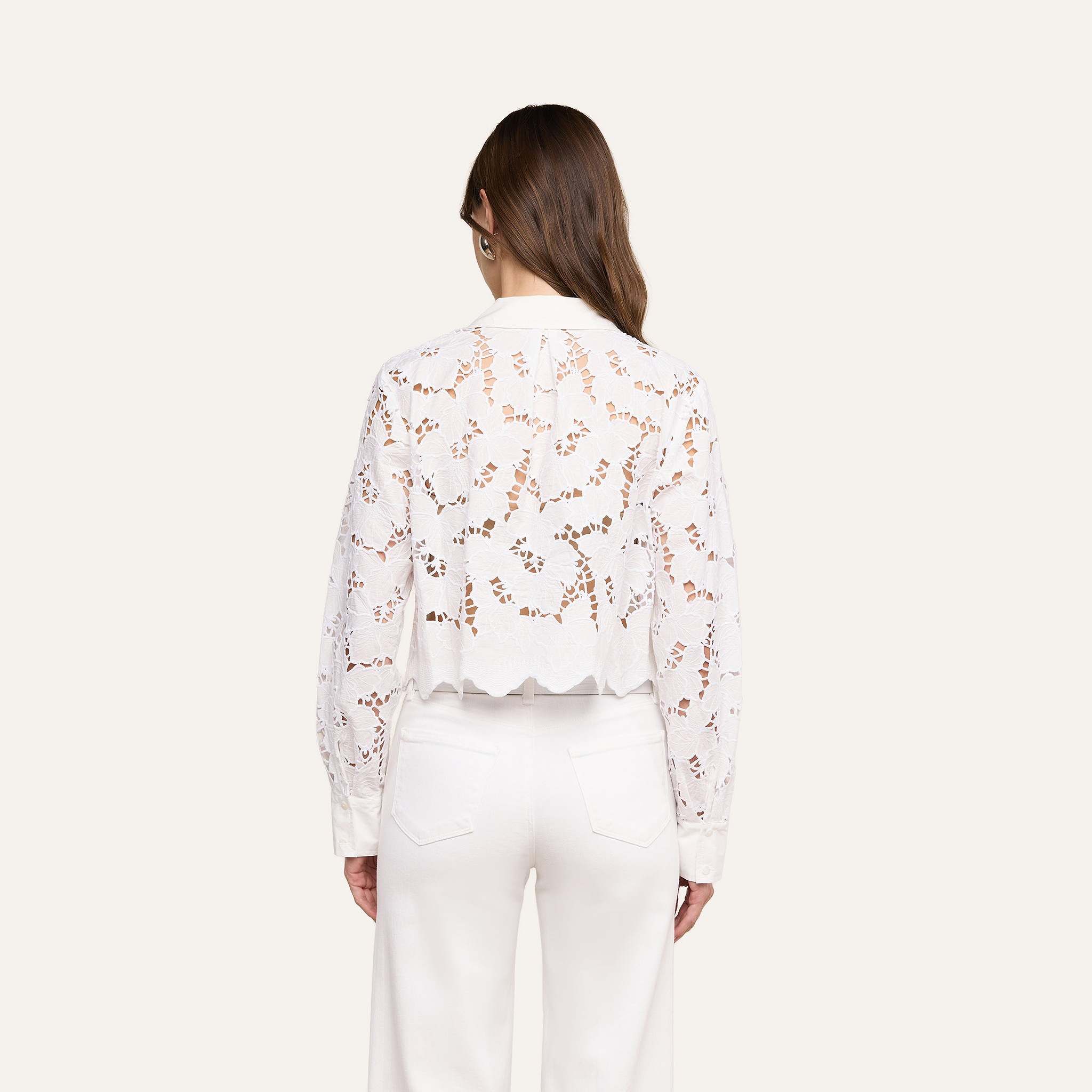 A woman stands with her back to the camera, wearing the Seychelle Cropped Eyelet Blouse—an all-white, long-sleeve top featuring a semi-sheer floral eyelet pattern and scalloped hem—paired with high-waisted white pants. Her brown hair is worn down, and the background is a plain light beige.