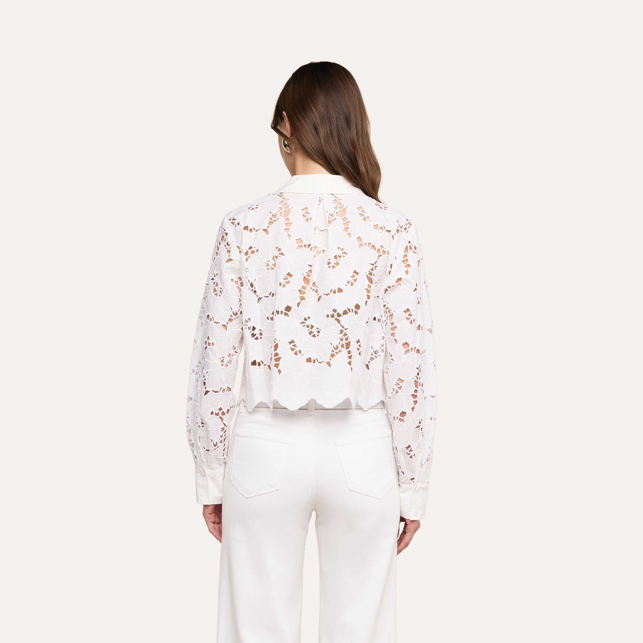 A woman stands with her back to the camera, wearing the Seychelle Cropped Eyelet Blouse—an all-white, long-sleeve top featuring a semi-sheer floral eyelet pattern and scalloped hem—paired with high-waisted white pants. Her brown hair is worn down, and the background is a plain light beige.