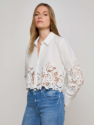 A woman with long blonde hair wears the L'AGENCE Seychelle Cropped Eyelet Blouse, a white button-down featuring floral eyelet lace cutouts at the hem and sleeves, paired with high-waisted blue jeans against a plain light background.